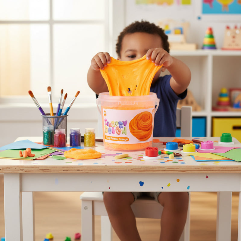 Child stretching bright fluoro orange sensory dough above a table filled with art supplies, paints, and creative classroom materials during sensory play.