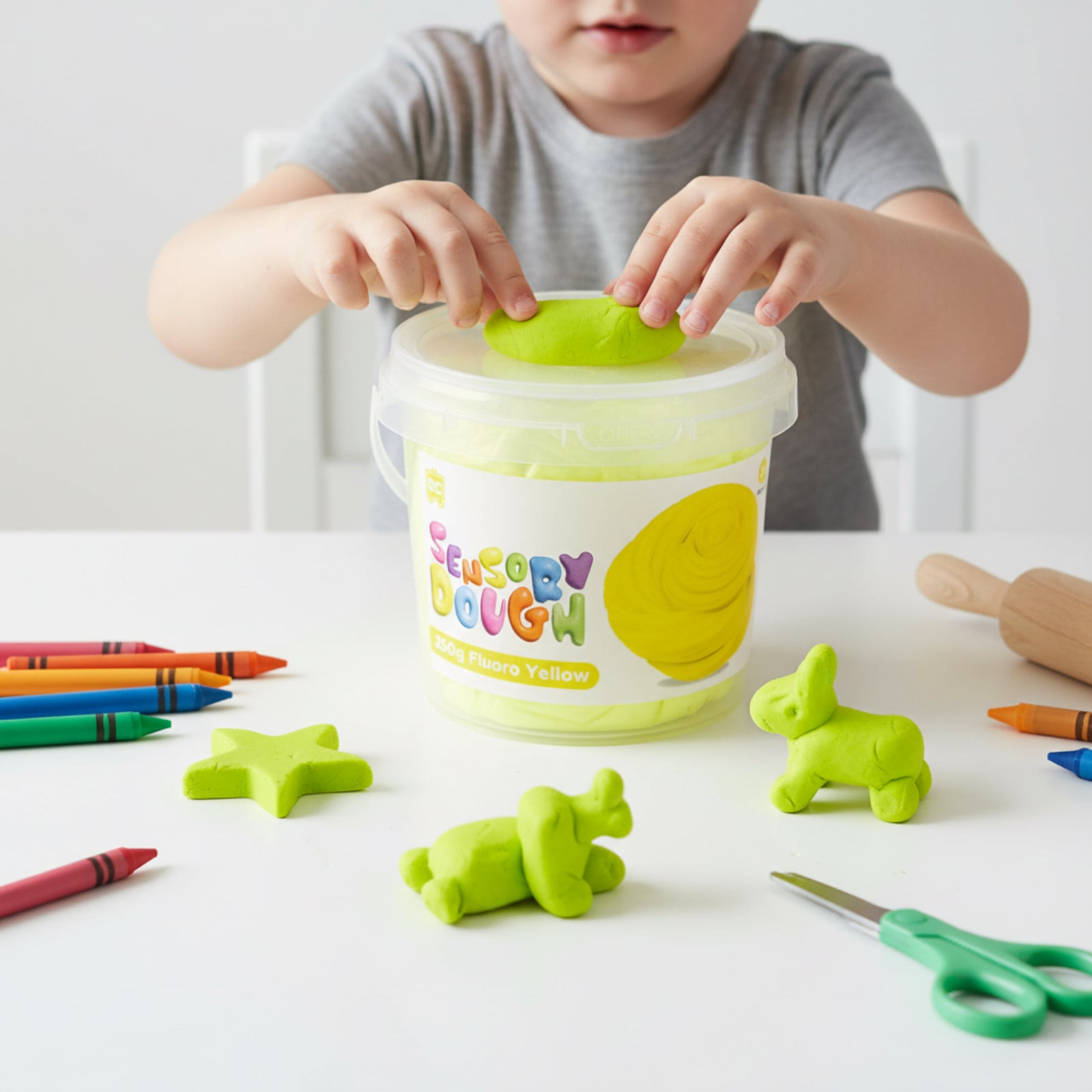 Young child placing fluoro yellow sensory dough into the reusable Educational Colours tub surrounded by shaped dough figures and art supplies.