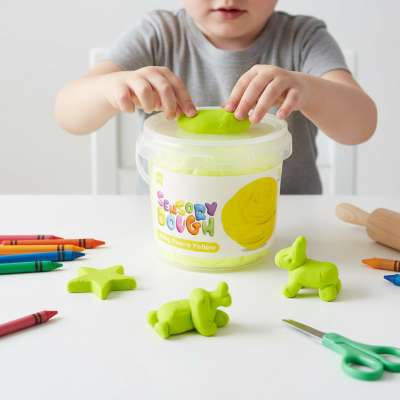 Young child placing fluoro yellow sensory dough into the reusable Educational Colours tub surrounded by shaped dough figures and art supplies.