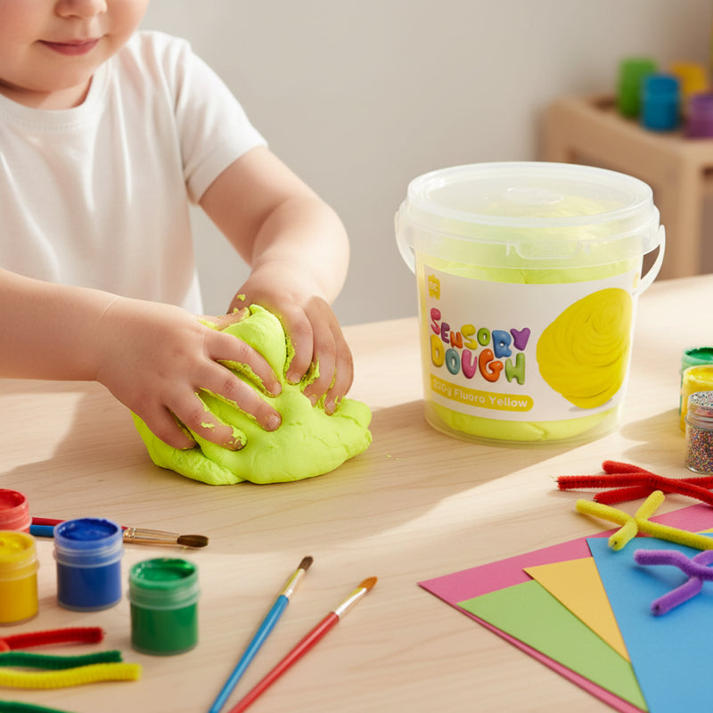 Child pressing and stretching bright fluoro yellow sensory dough beside colourful paints and craft materials during creative playtime.