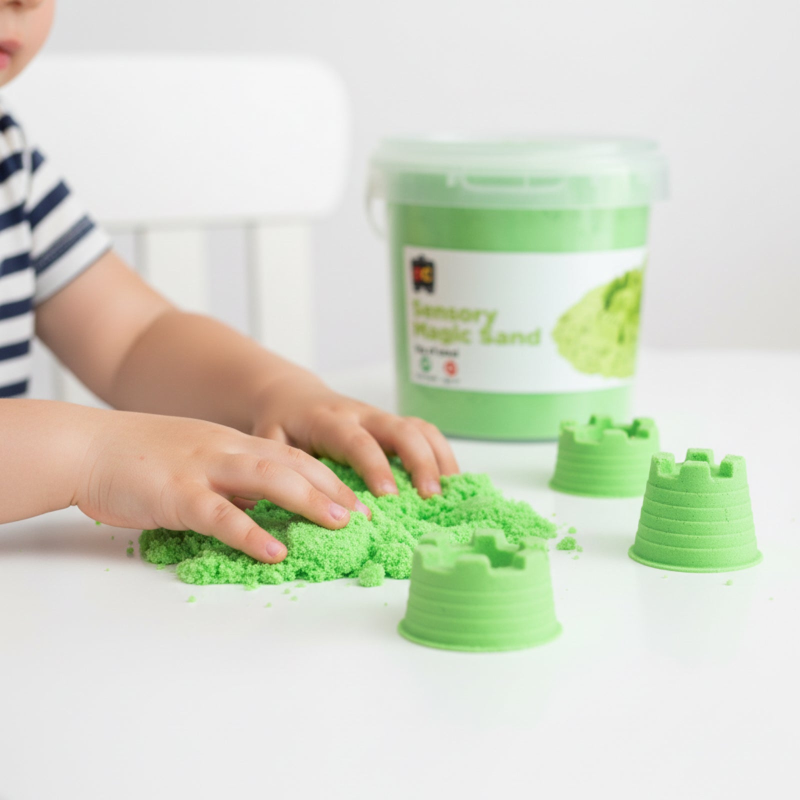 Young child exploring soft green sensory magic sand with castle moulds and reusable storage container during imaginative tactile play activity.