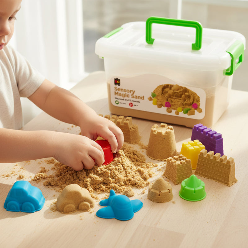 Young child pressing mould into natural sensory magic sand with castle walls, vehicle shapes, and assorted colourful moulds arranged on wooden table.
