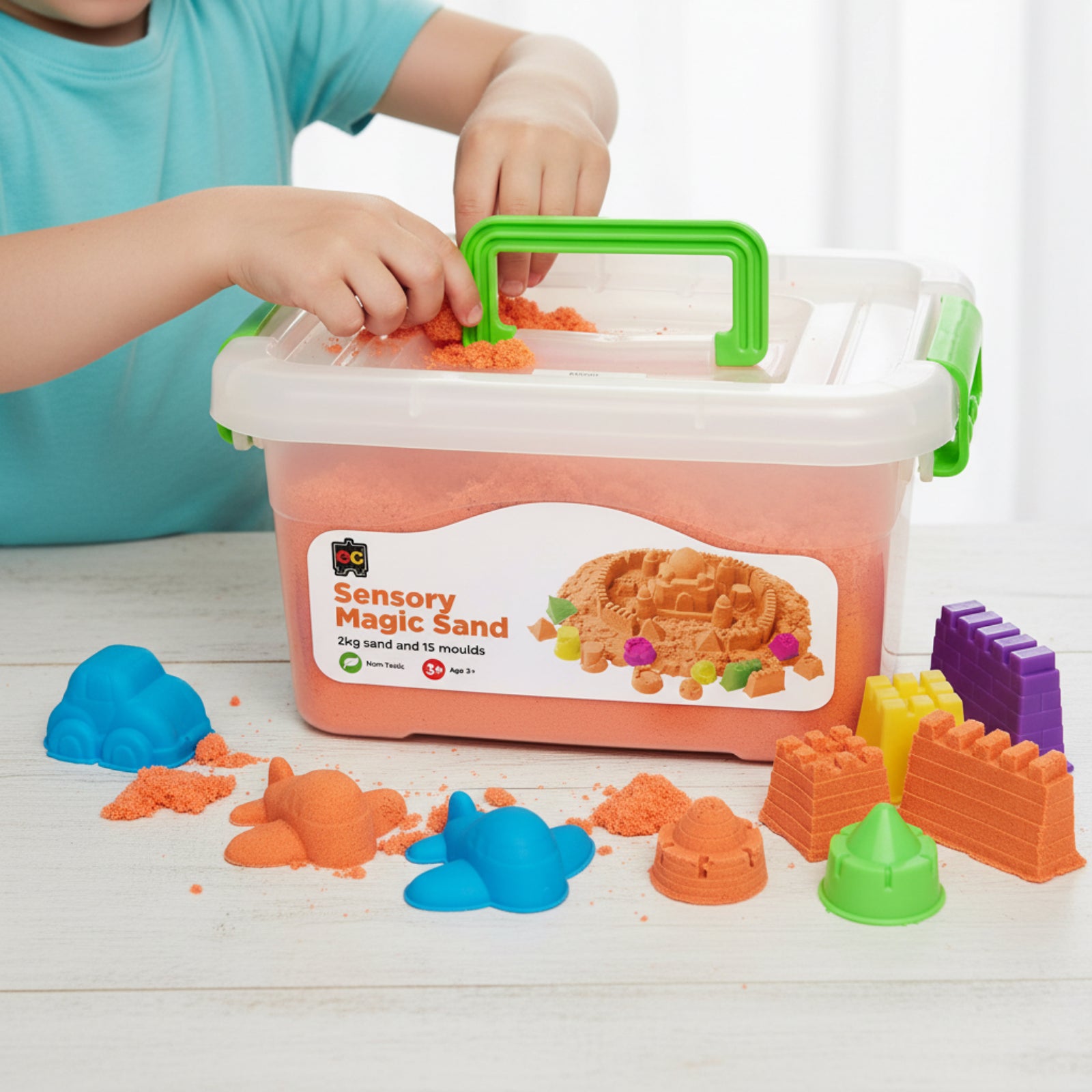 Young child placing orange sensory magic sand into reusable storage tub with colourful castle moulds and shaping accessories arranged on tabletop.