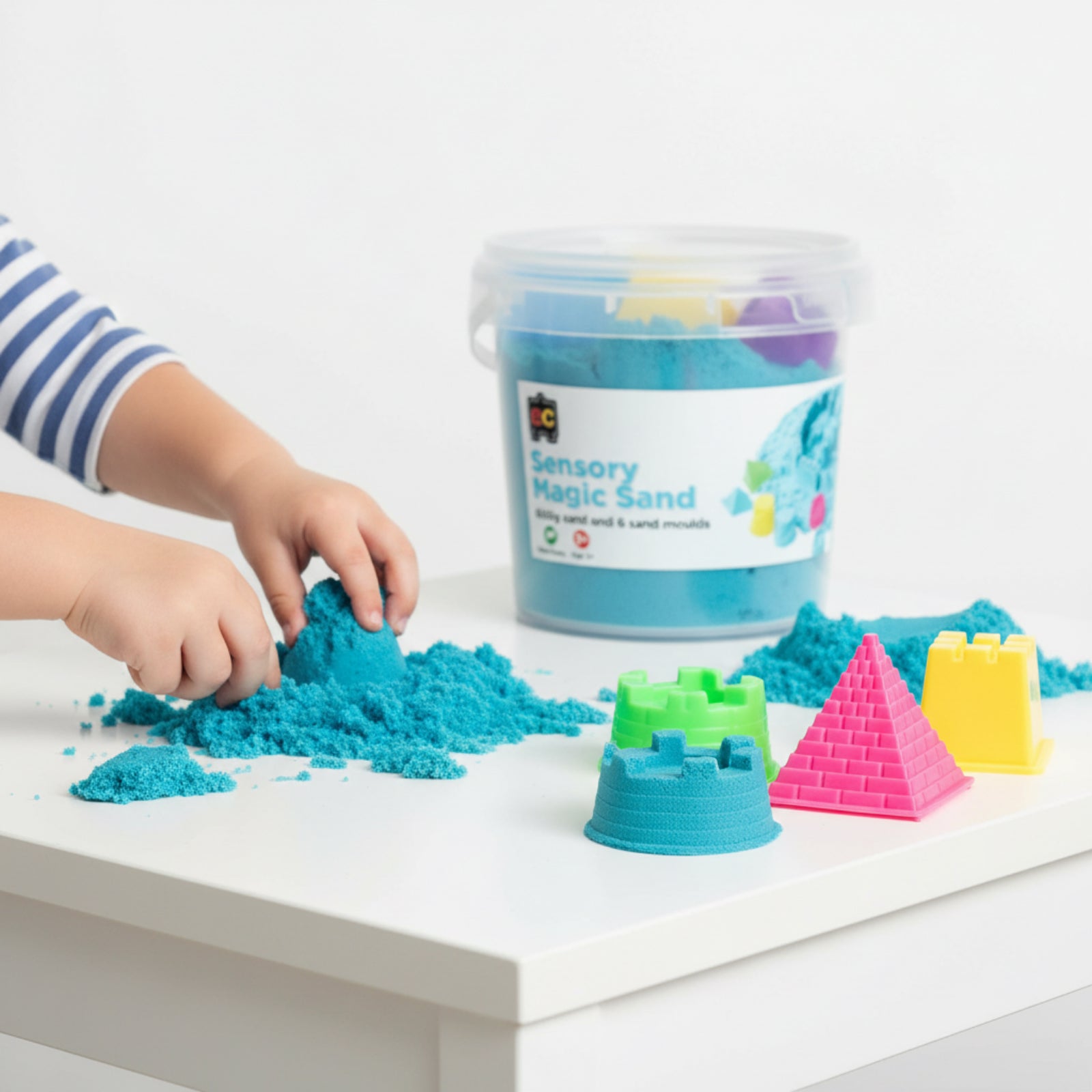 Young child using moulds with blue sensory magic sand on white play table surrounded by colourful castle and pyramid shaping tools.