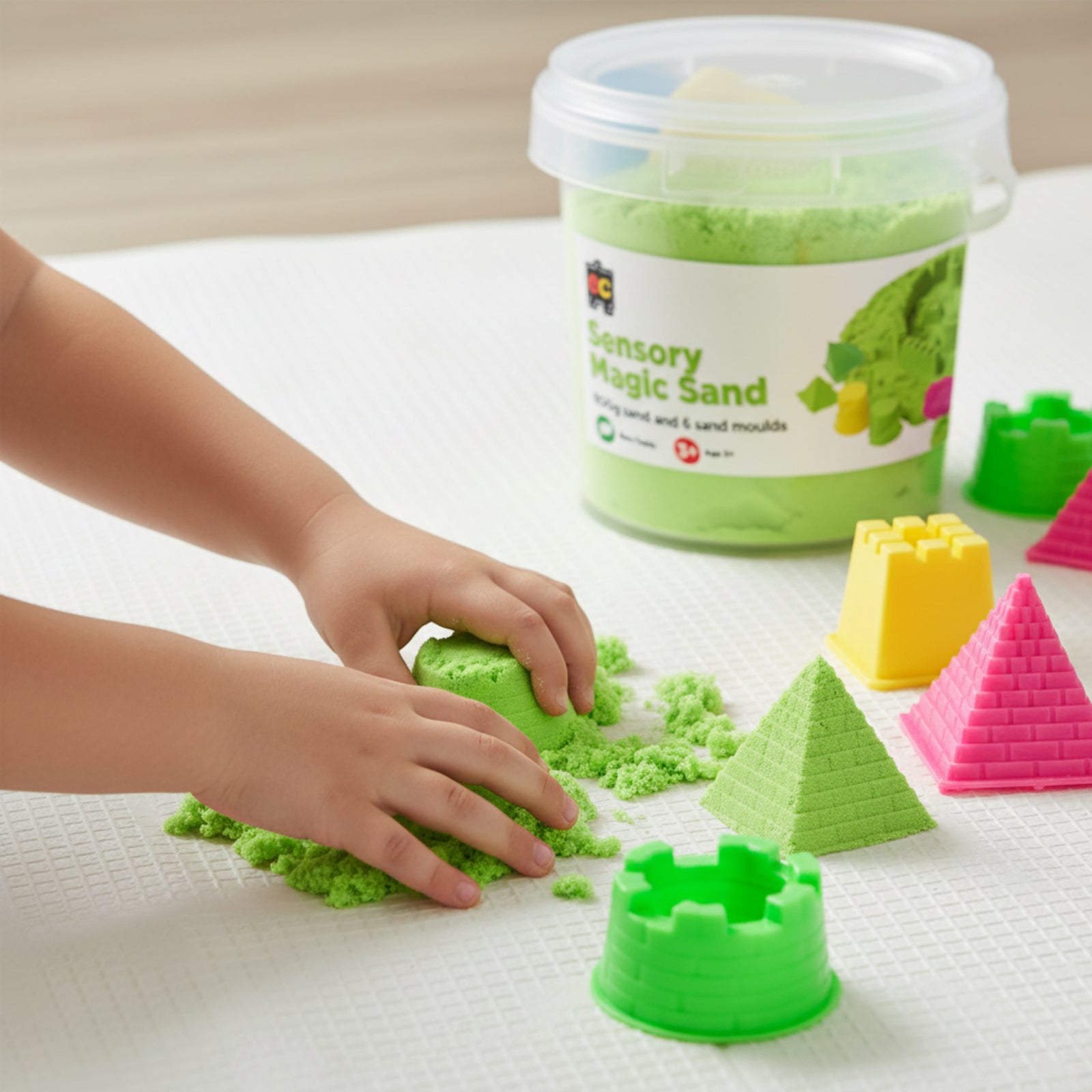 Child shaping bright green Educational Colours Sensory Magic Sand with moulds on textured play surface beside colourful pyramid and castle accessories.
