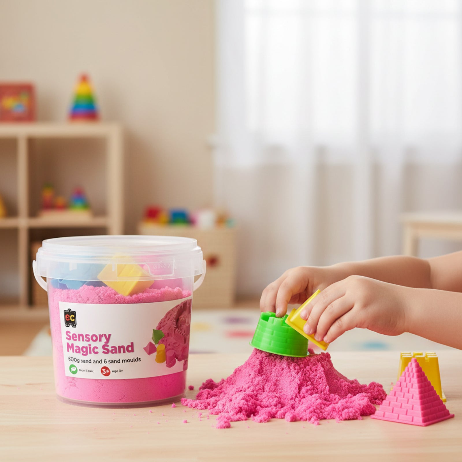 Young child creating detailed sand structures using pink sensory magic sand and colourful moulds in a warm indoor learning environment.
