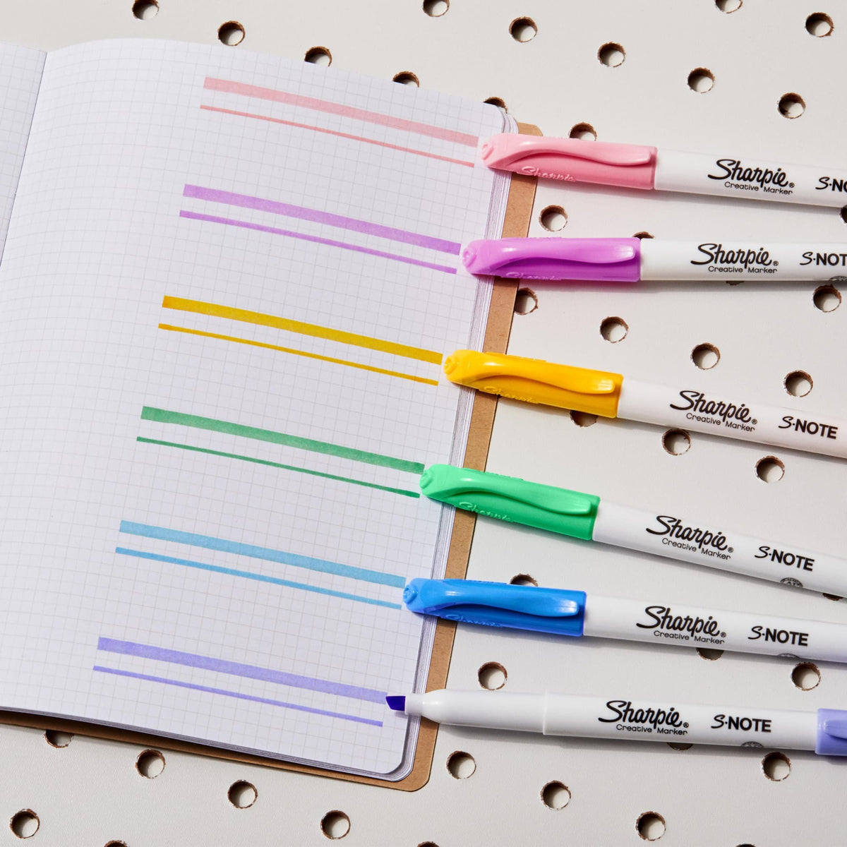 Six Sharpie S-Note markers in pink, purple, yellow, green, blue and lavender laid across a grid notebook with matching strokes