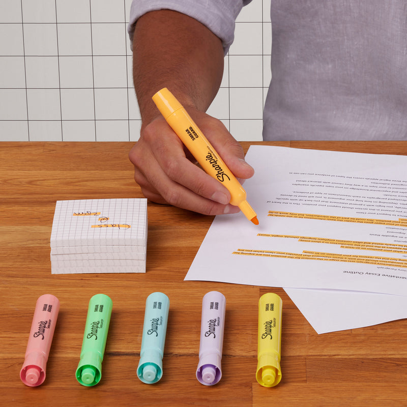 Person highlighting a printed document with a pastel orange Sharpie Tank highlighter, with pink, green, blue, purple and yellow pastels on the desk
