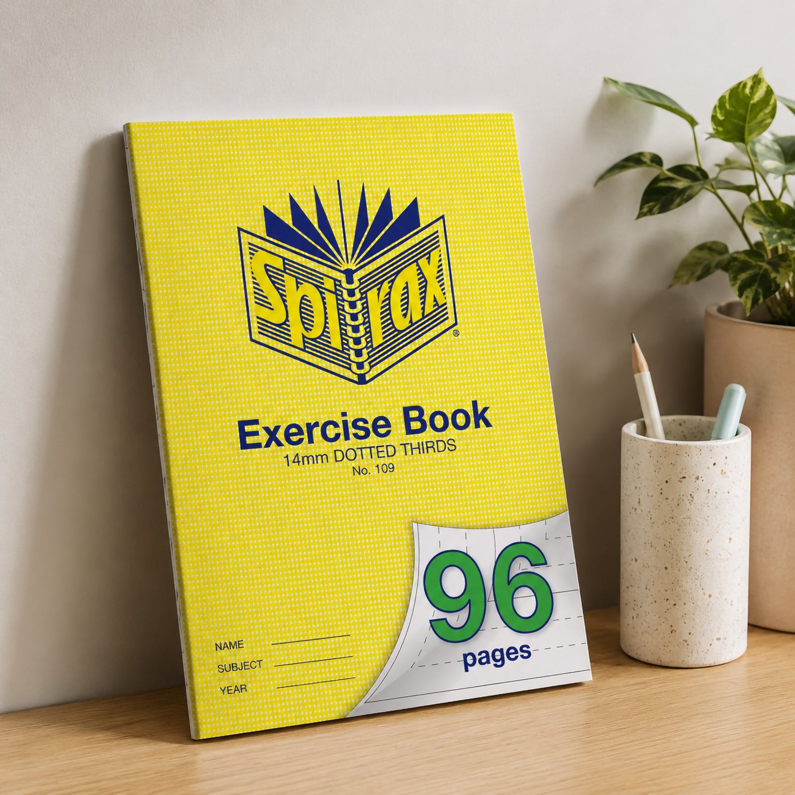 Spirax 109 A4 Exercise Book standing upright against a neutral wall on a wooden desk with stationery holder and indoor plant, creating a modern classroom or home office lifestyle setting.
