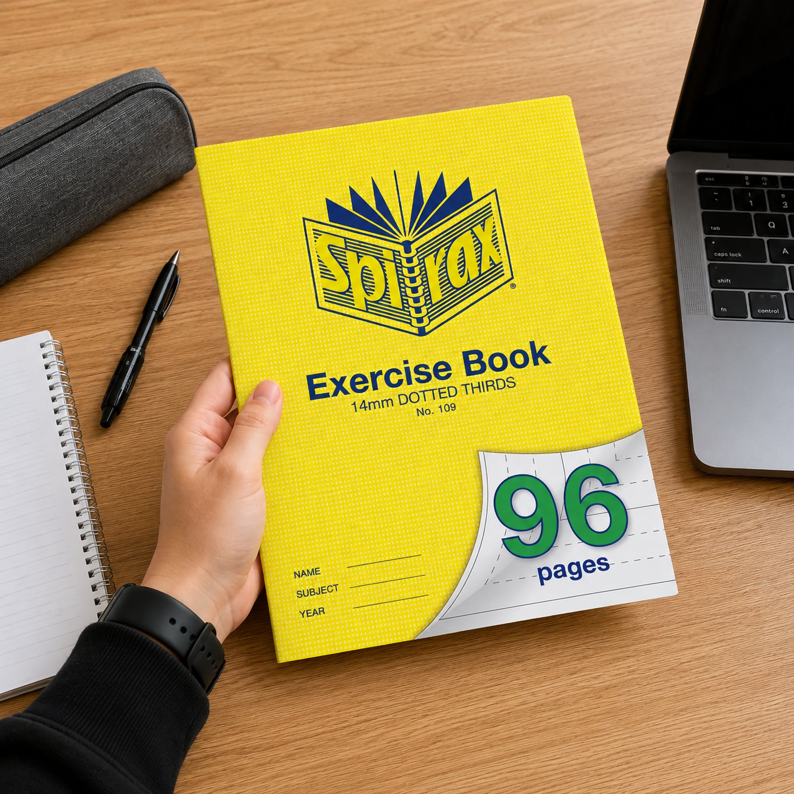 Student holding a bright yellow Spirax 109 A4 Exercise Book with 96 pages and 14mm dotted thirds ruling over a wooden study desk beside a laptop, notebook, pen and pencil case, highlighting everyday classroom and home study use.