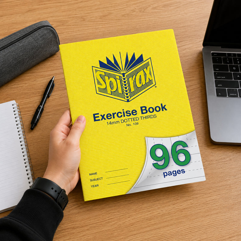 Student holding a bright yellow Spirax 109 A4 Exercise Book with 96 pages and 14mm dotted thirds ruling over a wooden study desk beside a laptop, notebook, pen and pencil case, highlighting everyday classroom and home study use.