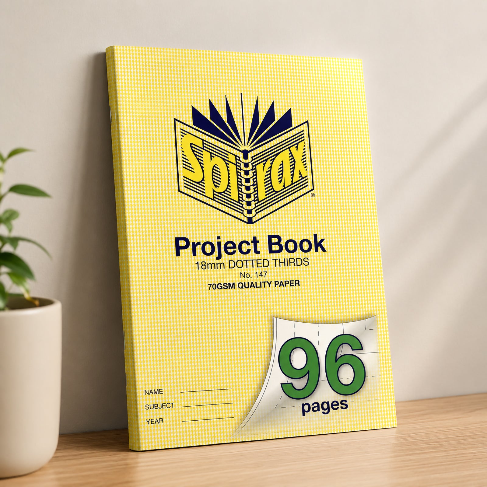 Spirax 147 A4 Project Book standing upright against a neutral wall on a wooden desk with a potted plant, showcasing the bright yellow cover in a clean classroom or home study environment.