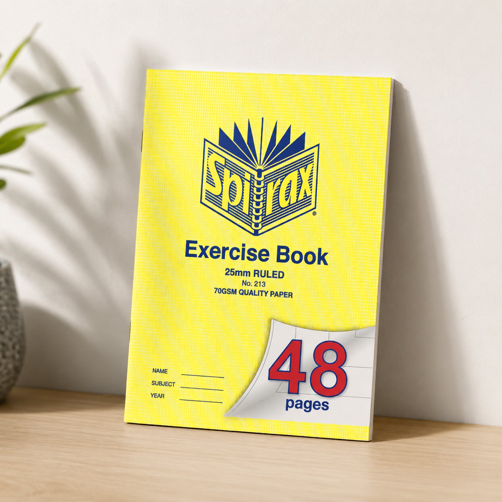 Spirax 213 A4 Exercise Book standing upright against a neutral wall on a wooden desk, surrounded by simple classroom stationery and greenery, highlighting the wide 25mm ruled design for foundation students.