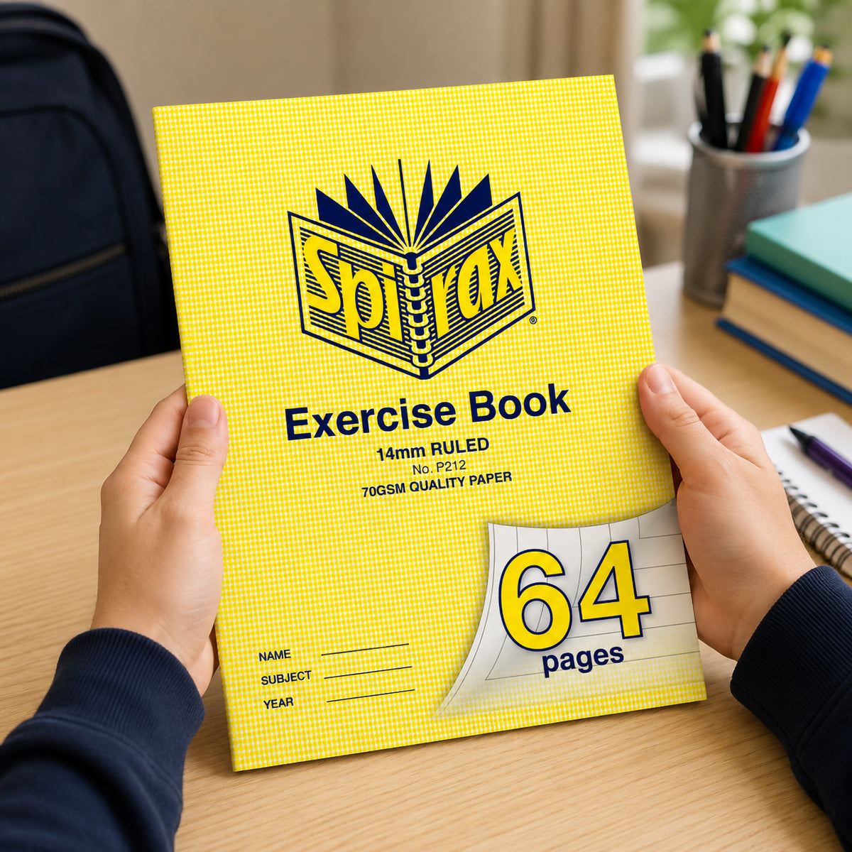 Two hands holding a Spirax P212 A4 exercise book with the yellow 64 pages corner showing, pencils and books behind