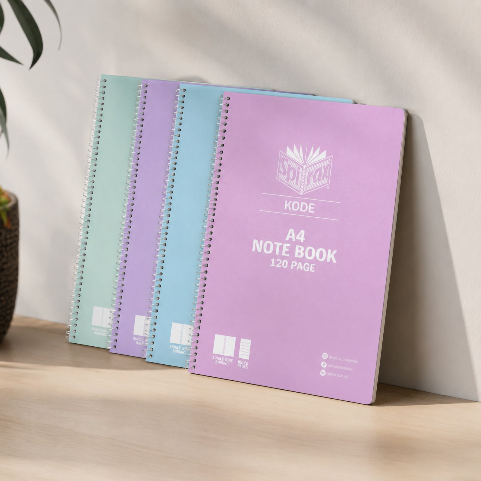 Spirax Kode A4 metallic notebooks standing against a neutral wall on a wooden desk, showing the pastel pink, blue, purple and mint covers in a clean study or office setting.