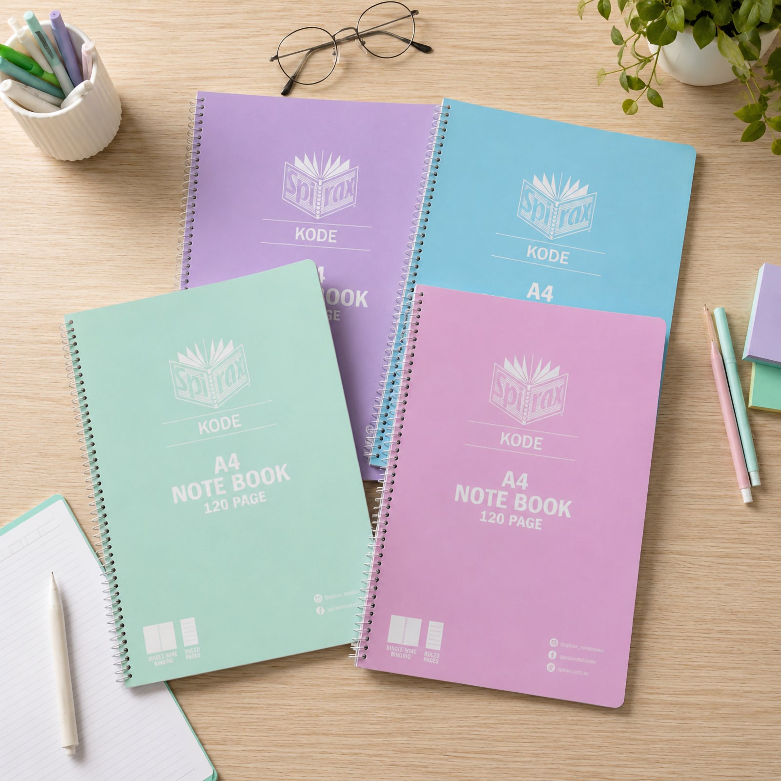 Top view of four Spirax Kode A4 metallic notebooks arranged across a desk with glasses, pens, plant and note paper, highlighting the assorted pastel covers and single wire binding.
