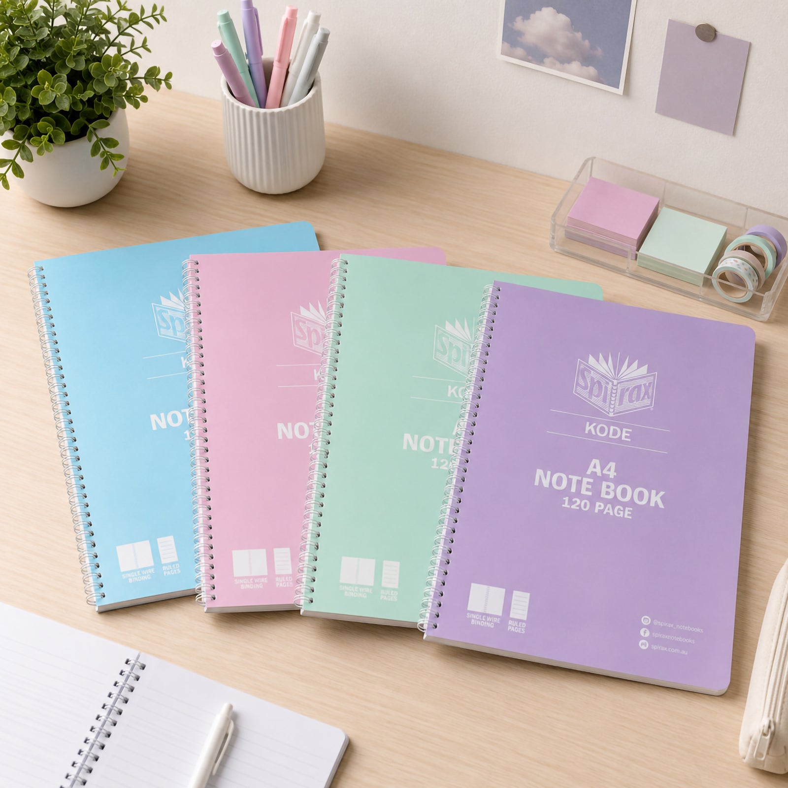Assorted Spirax Kode A4 metallic notebooks in pastel blue, pink, mint and purple displayed on a wooden study desk with pens, sticky notes, plant and stationery, showing a bright organised workspace for school or study.