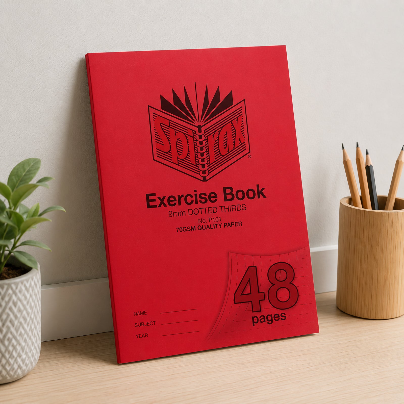 Spirax P101 A4 Exercise Book leaning against a neutral wall on a clean timber desk with potted plants and pencils, presenting the durable classroom design in a bright learning space.