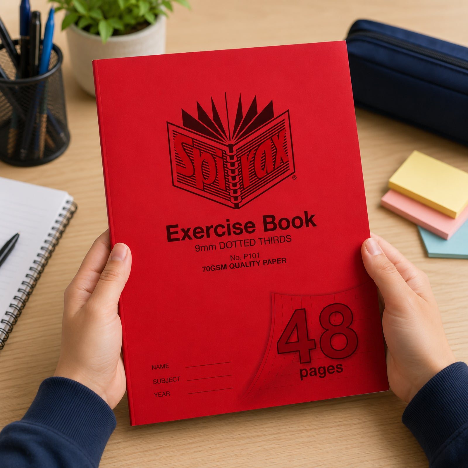 Hands holding a Spirax P101 A4 Exercise Book with a bright red cover on a tidy study desk surrounded by notebooks, stationery, sticky notes and a pencil case, highlighting the 48 page format and 9mm dotted thirds ruling designed for handwriting practice.