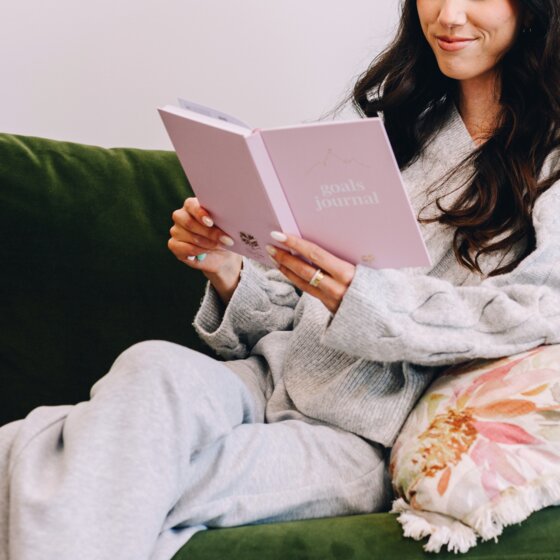 Woman relaxing on a green sofa reading a blush Spirax Platinum goals journal held open in both hands