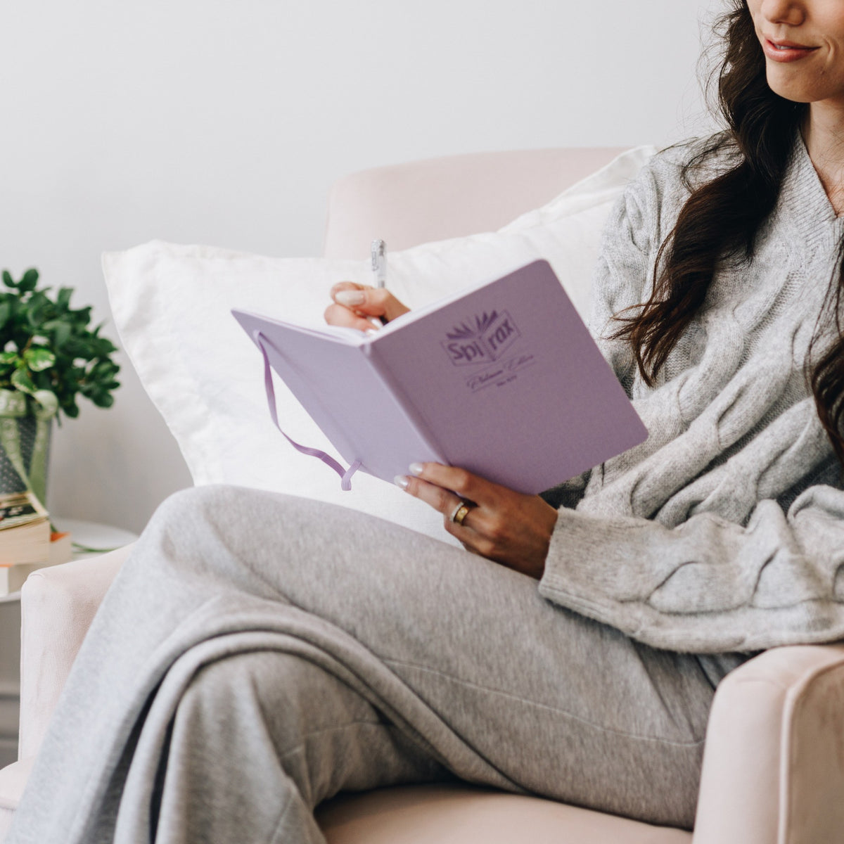 Woman relaxing on a sofa with a grey throw over her legs, writing in a lilac Spirax Platinum linen-look journal held open on her lap