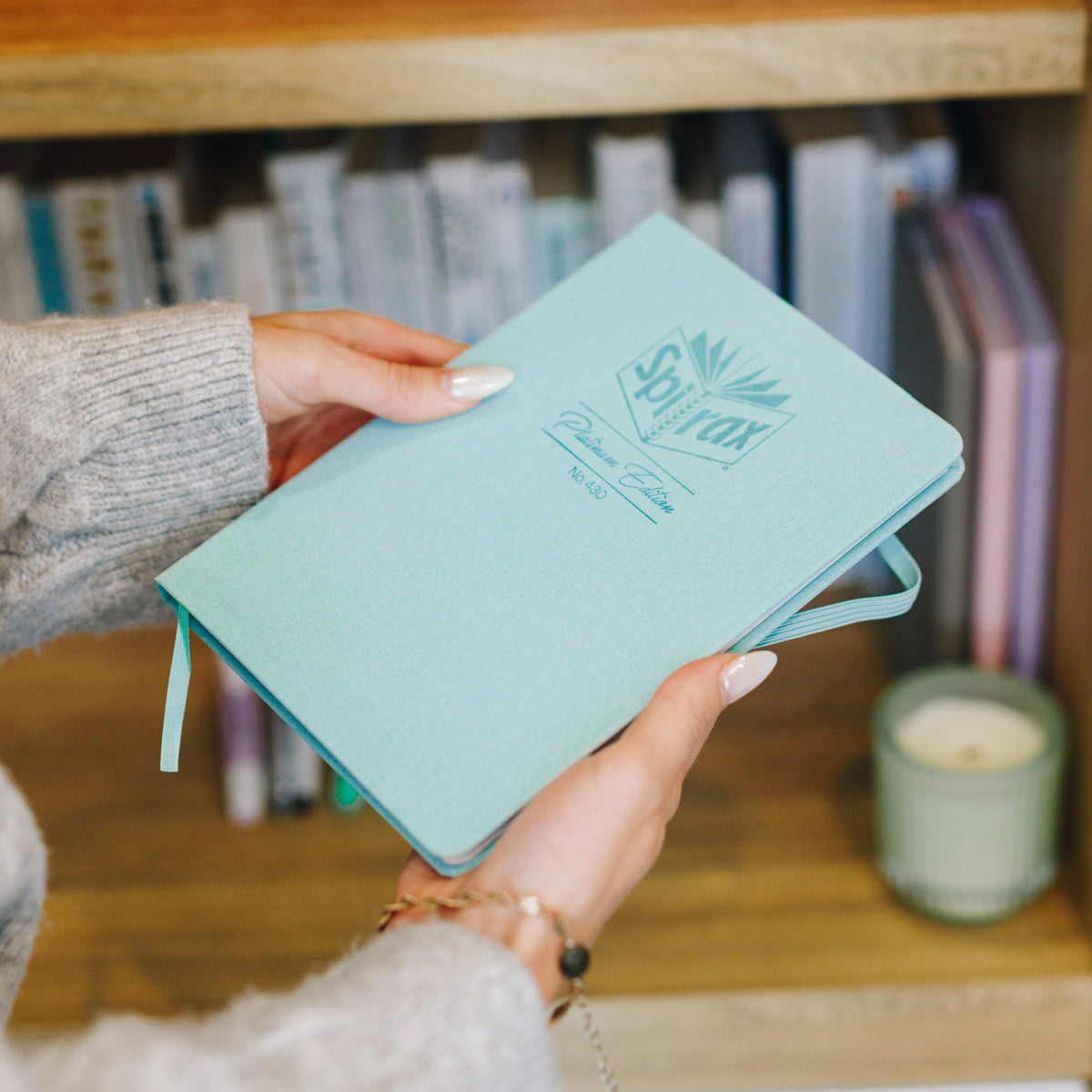 Hands holding a Spirax Platinum Edition No. 430 journal with a sage linen-look cover and matching elastic strap, in front of a timber bookshelf and a candle