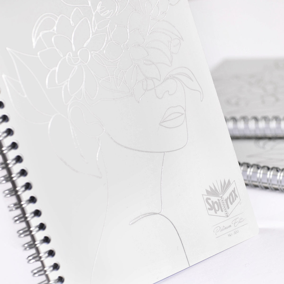 Close view of the white Spirax Platinum Edition No. 404 notebook cover with a raised line drawing of a face framed by flowers, beside its spiral binding