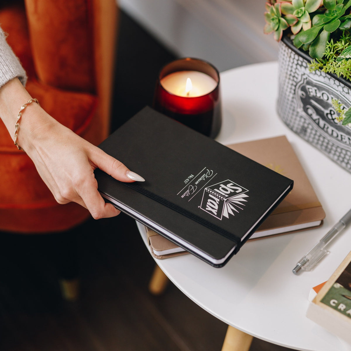 Hand placing a Spirax Platinum Edition No. 437 journal in jet black faux leather with silver foil on a small side table beside a red candle, a potted plant and a latte journal