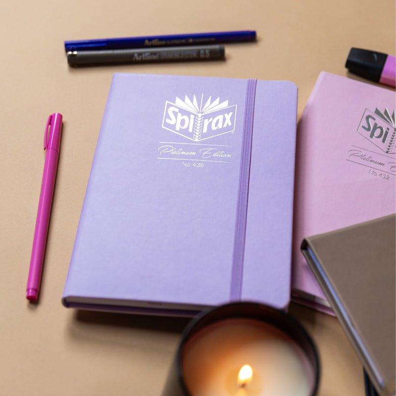 Spirax Platinum Edition No. 436 journal in lilac faux leather with a silver foil logo and lilac elastic strap, on a desk beside felt pens, a candle and a rose journal