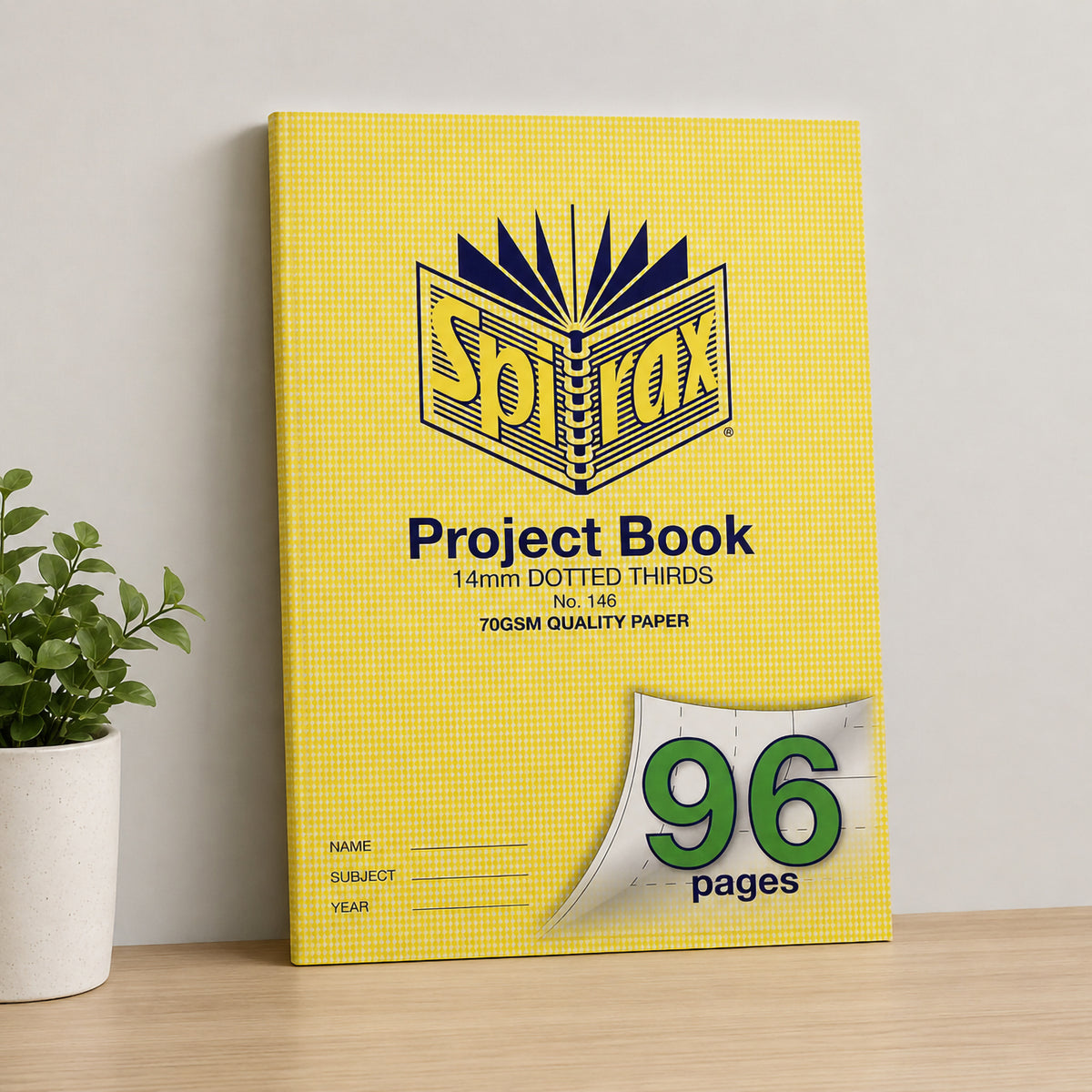 Spirax 146 project book standing against a pale wall on a wooden desk beside a potted plant, yellow cover facing forward