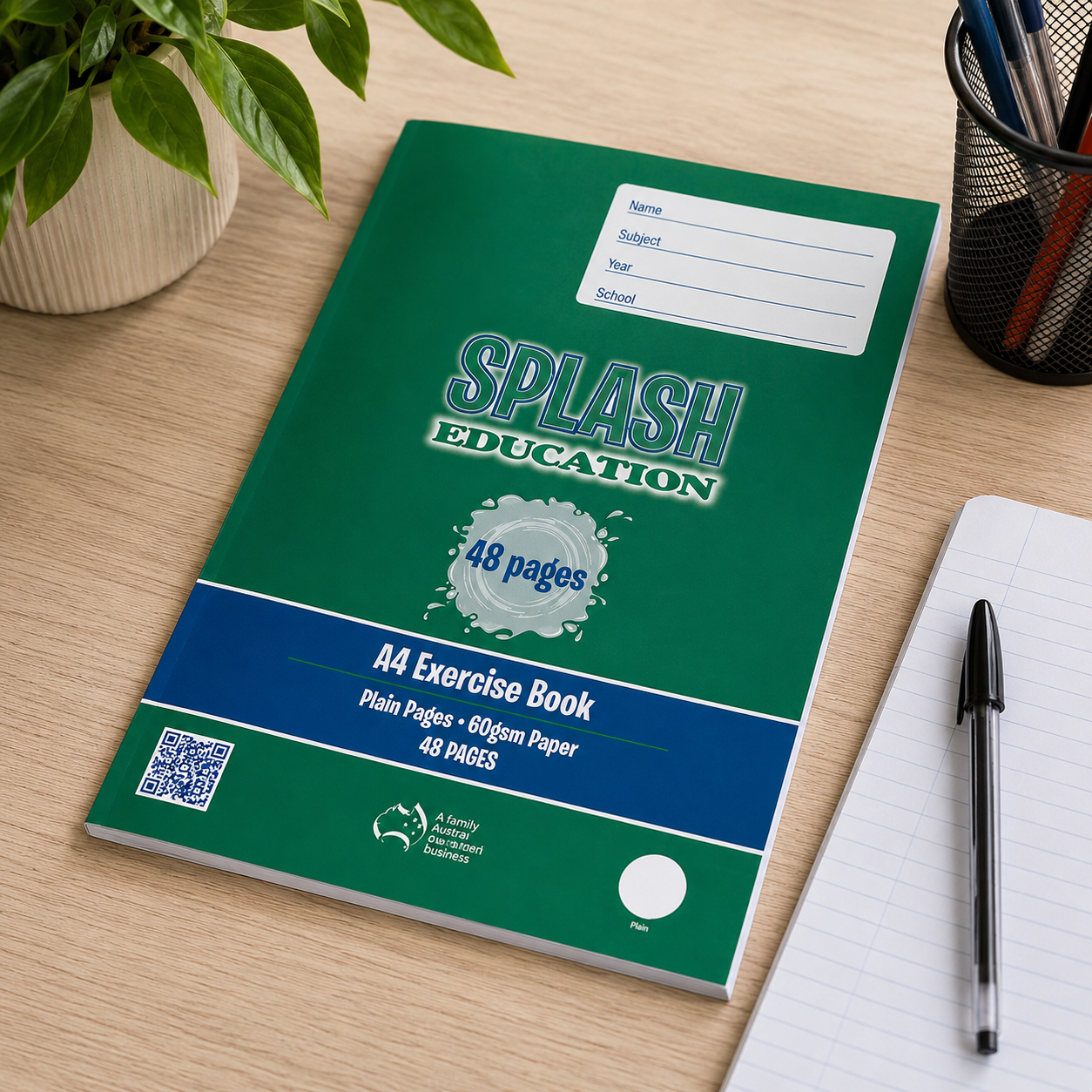 Splash A4 plain exercise book on a tidy classroom desk with stationery, plant and notebook nearby.