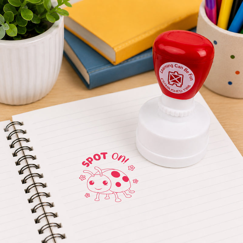 Classroom desktop scene featuring the Spot On self inking stamp standing on an open notebook. The bright ladybird themed design creates a fun and memorable reward that encourages confidence and motivates young learners.