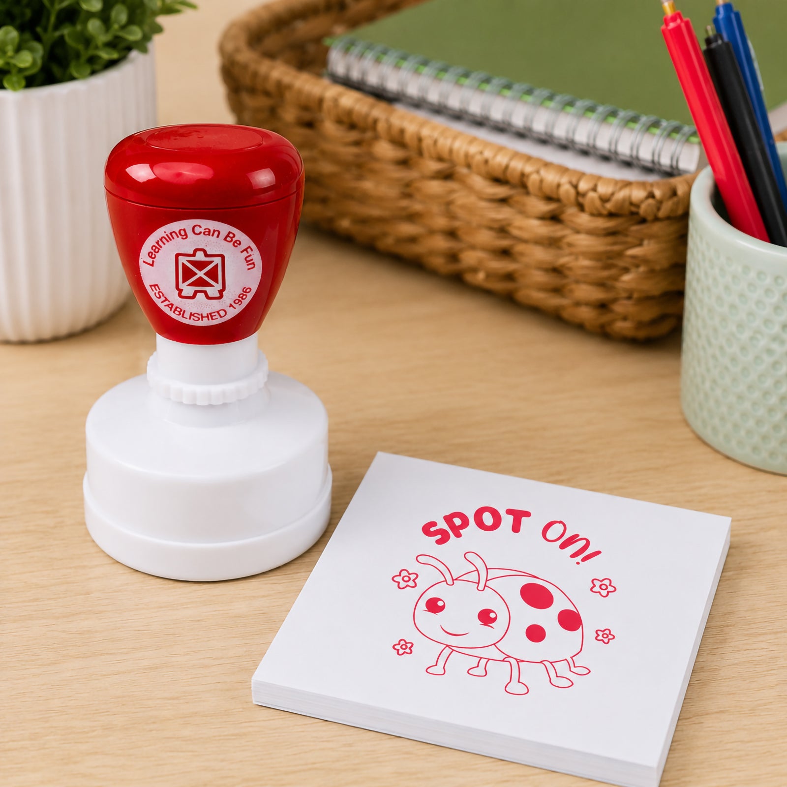 Red Spot On merit stamp positioned next to a stack of note paper in a learning environment. The friendly ladybird design and bold red impression provide an engaging way for teachers to reward effort, participation and completed classroom work.