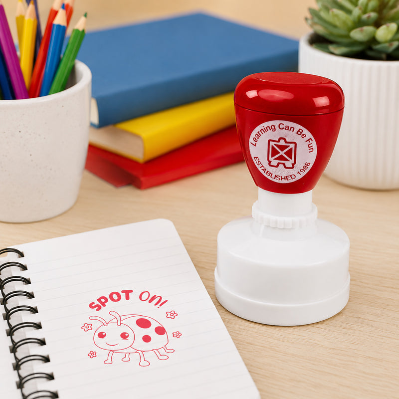 Learning Can Be Fun Spot On Merit Stamp displayed beside a notebook on a classroom desk. The self inking reward stamp creates a cheerful red ladybird impression with the Spot On! message, making it ideal for recognising student achievement and positive learning outcomes.