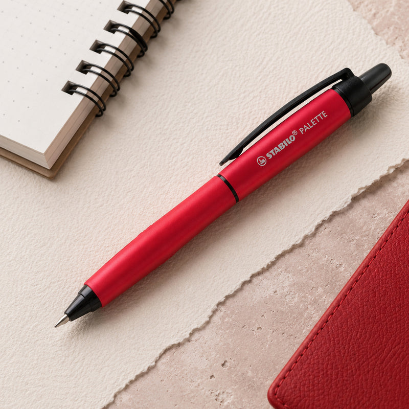 Top view of the STABILO Palette Retractable Rollerball Gel Pen Red resting beside a spiral notebook and matching red planner. The sleek metallic barrel and ergonomic grip are highlighted in a clean workspace designed for professional note taking.