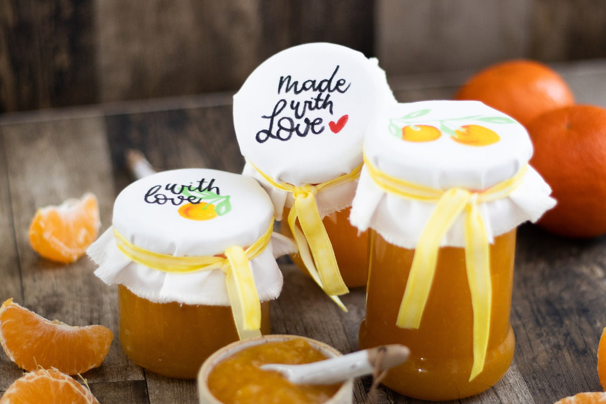 Glass jars of orange jam with fabric covers lettered made with love, tied with yellow ribbon, beside mandarins
