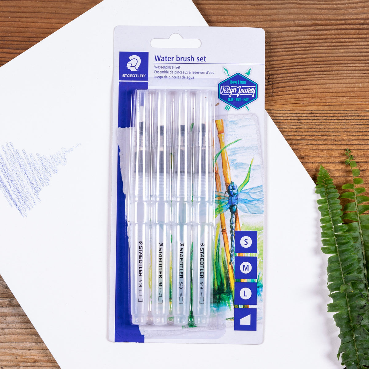 Staedtler photo of the 949 water brush set on white paper on a timber table with a fern and blue pencil scribble