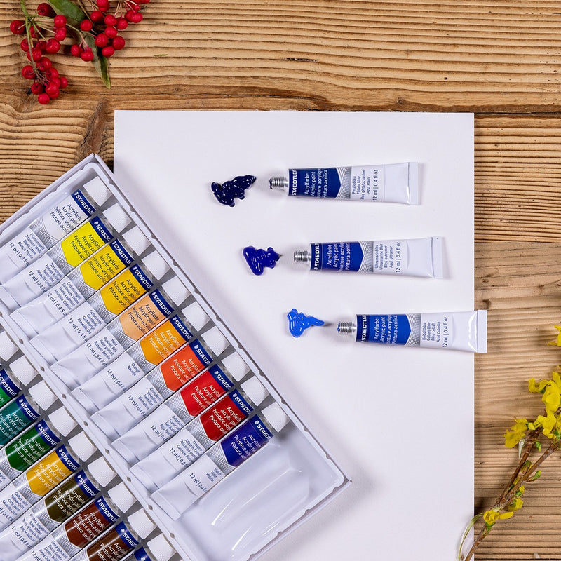 Open tray of Staedtler acrylic paint tubes on a wooden table beside a white board with blue paint squeezed out