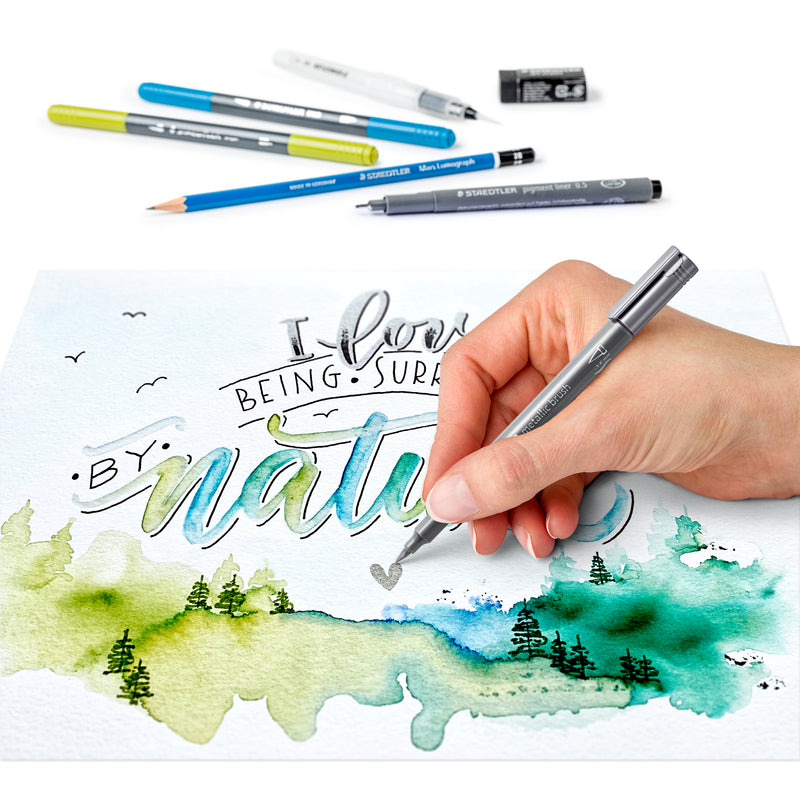 Hand outlining the word nature in grey over a blue and green watercolour landscape, with the brush pens, pencil and water brush above