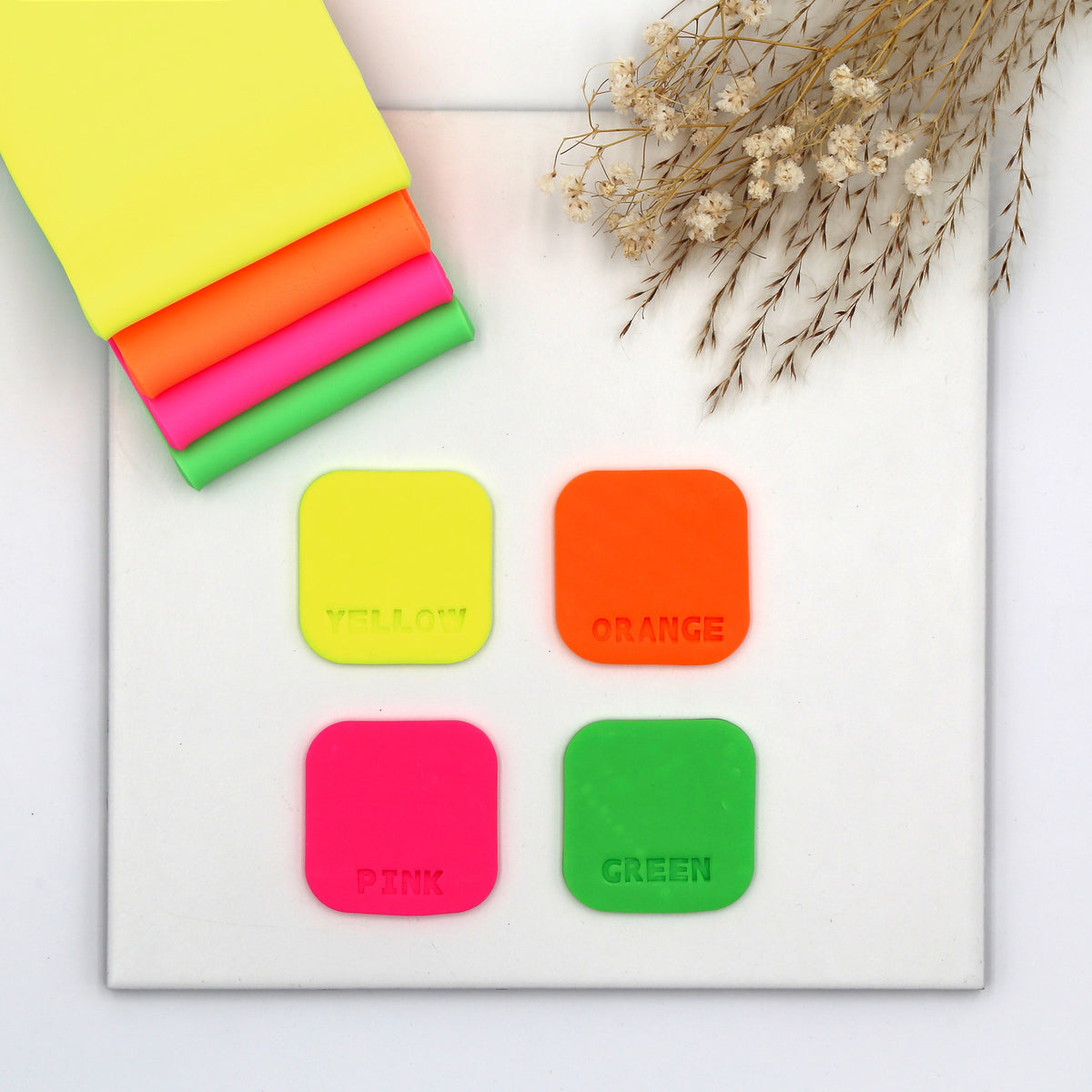 Four FIMO effect neon tiles in yellow, orange, pink and green beside folded sheets of neon clay