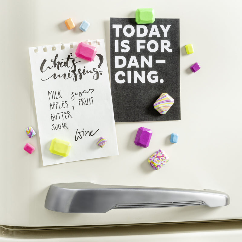 Bright neon clay magnets pinning a shopping list and a black card to a cream fridge door