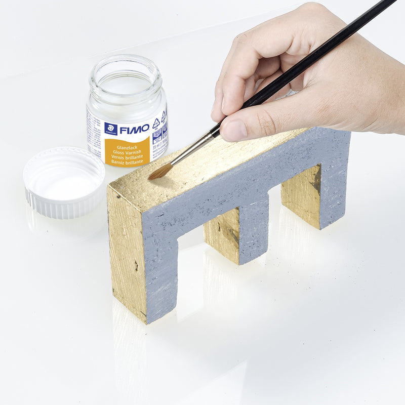 Hand brushing FIMO gloss varnish onto a gold and grey modelled piece, with the open jar and white lid beside it