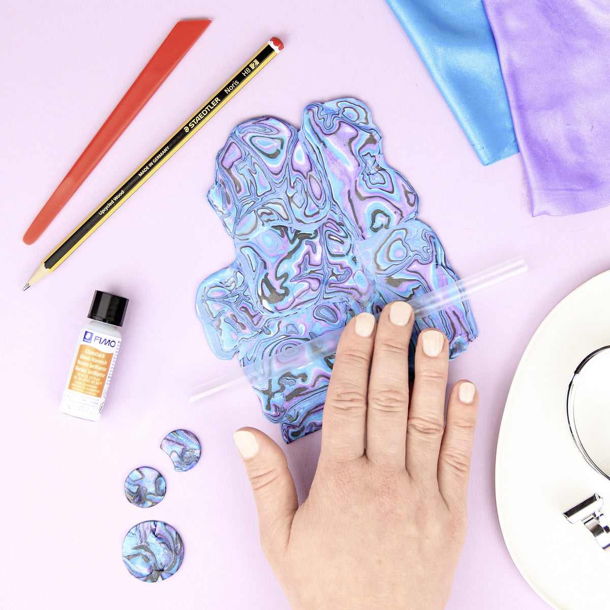 Hand smoothing a marbled turquoise, lilac and grey abalone-effect clay sheet next to round cut pieces and the varnish bottle