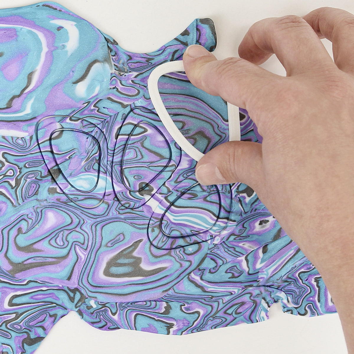 Hand pressing an oval cutter outline into a marbled abalone-effect clay sheet in turquoise, lilac and grey