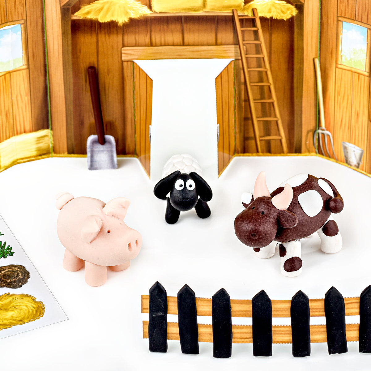 Finished clay pig, black faced sheep and brown spotted cow standing in the cardboard barn scene behind a fence