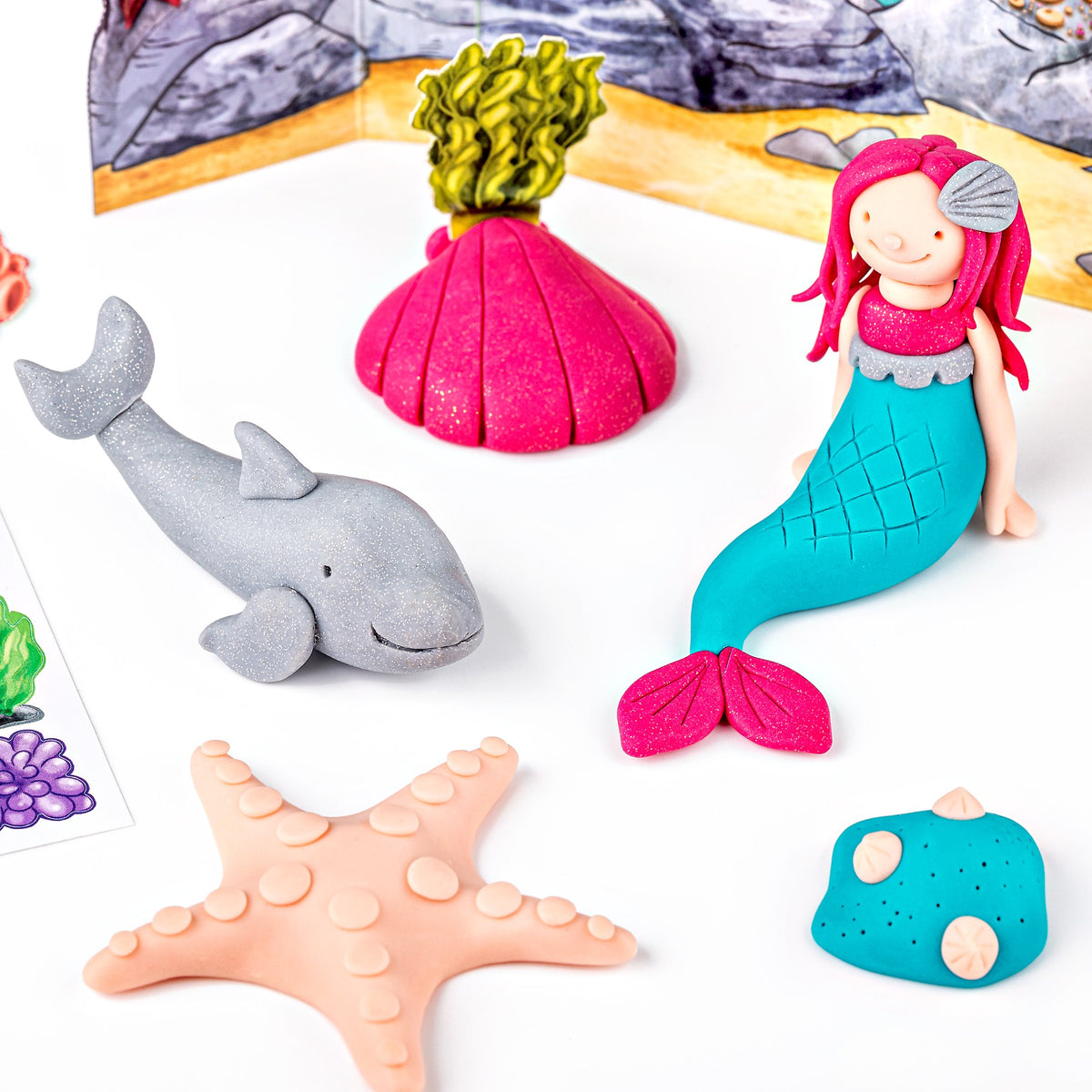 Clay mermaid, grey glitter dolphin, pink shell, starfish and turquoise rock arranged on the reef scene