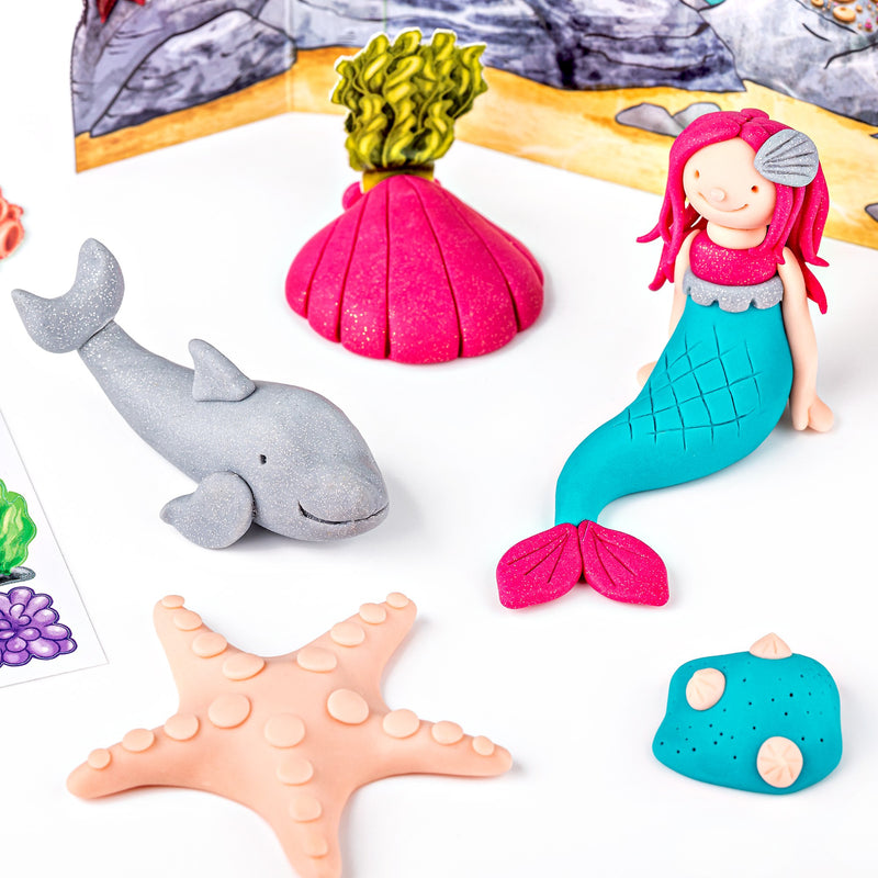 Clay mermaid, grey glitter dolphin, pink shell, starfish and turquoise rock arranged on the reef scene