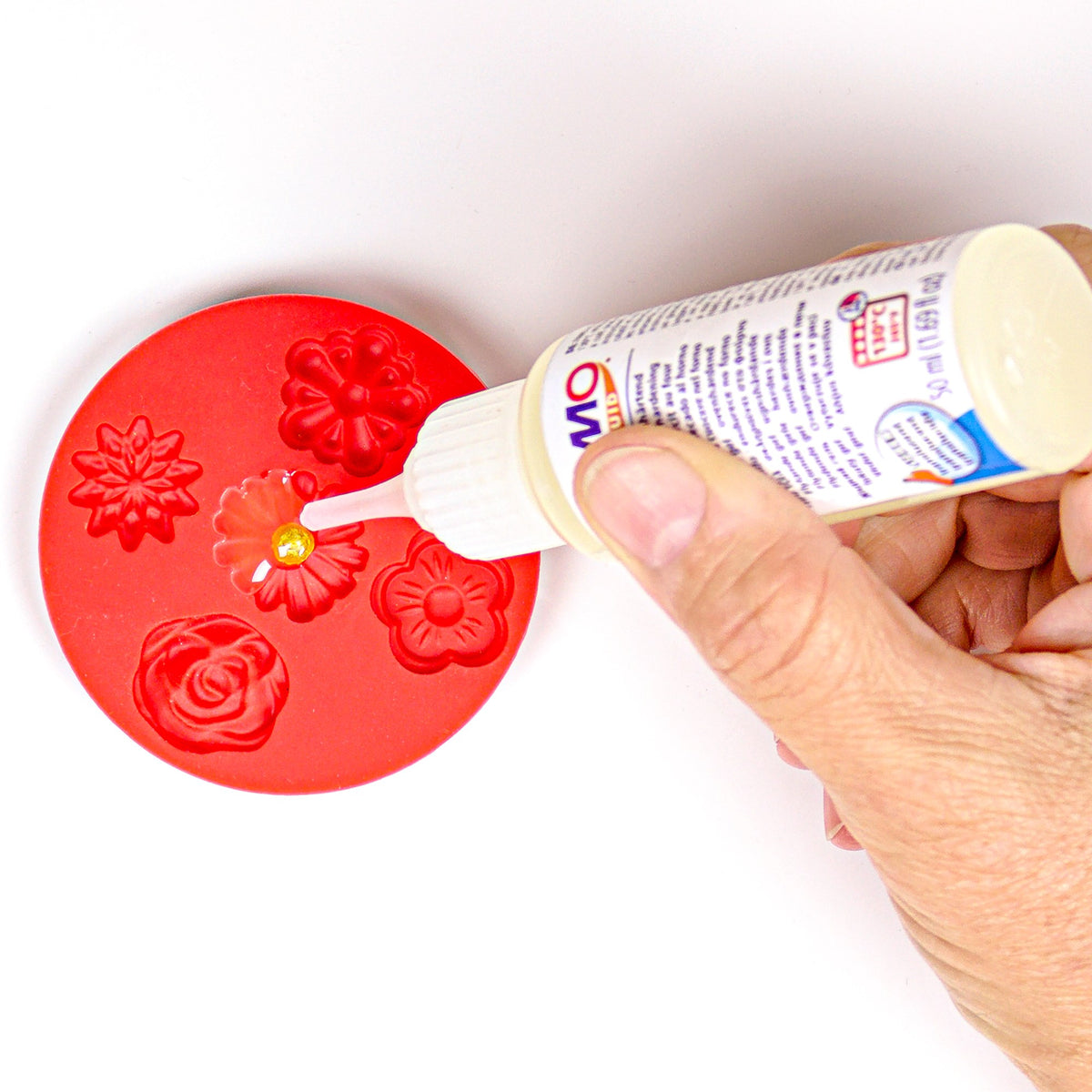 Hand squeezing a translucent FIMO liquid bottle onto red clay flowers in a mould, a range photo showing how the dosage tip places the gel