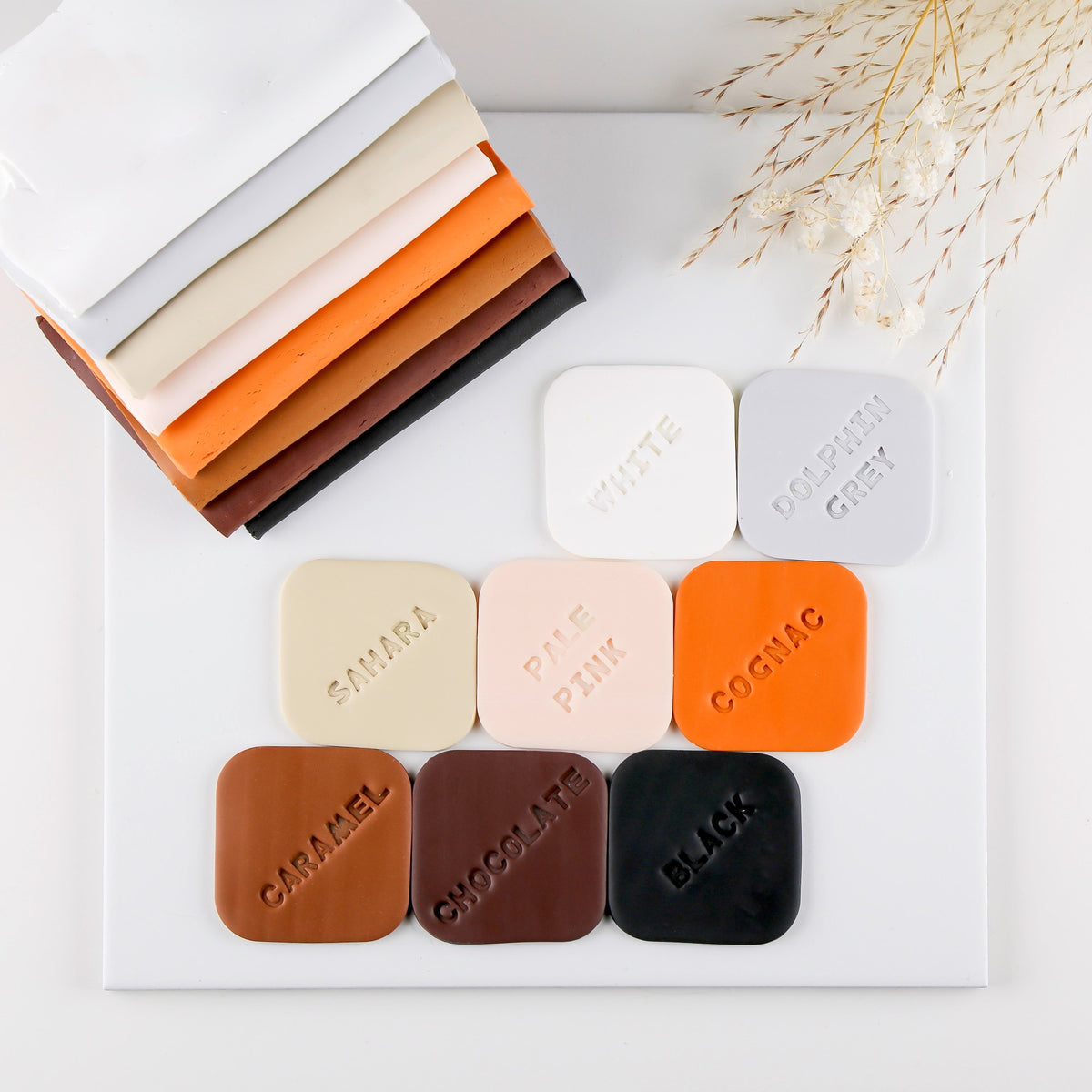 FIMO soft colour samples on a white tile in neutral shades with their names pressed in, including pale pink and black, beside stacked clay sheets