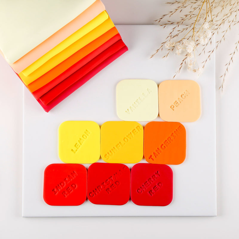 Warm FIMO soft colour samples on a white tile with names pressed in, from lemon and sunflower to tangerine and reds, beside stacked clay sheets